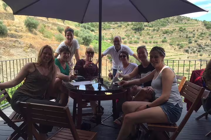 Group enjoying wine tasting under a shaded terrace in Douro Valley with vineyards in the background.