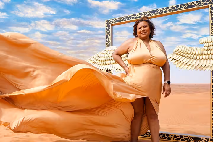 Woman in flowing golden dress poses in Dubai desert with decorative frame and wings, part of Flying Dress Photography Tour.