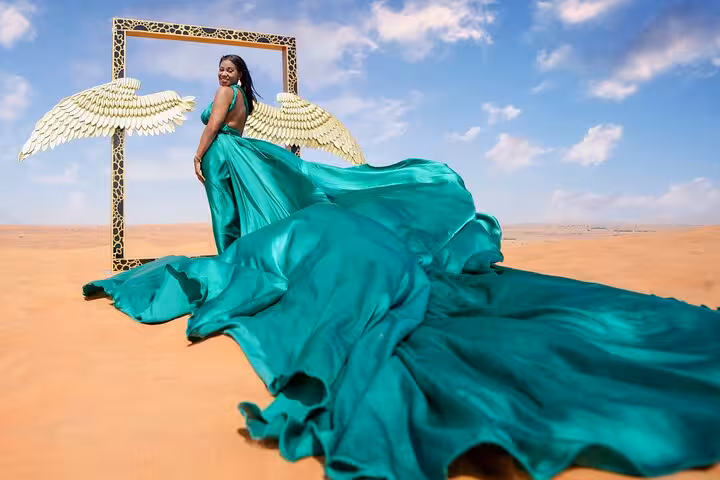 Woman in a flowing green dress poses with angel wings in the vast Dubai desert under a bright blue sky.