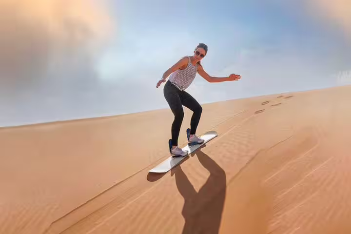 Adventurer sandboarding down a golden dune under a clear sky during a private morning Dubai desert safari.