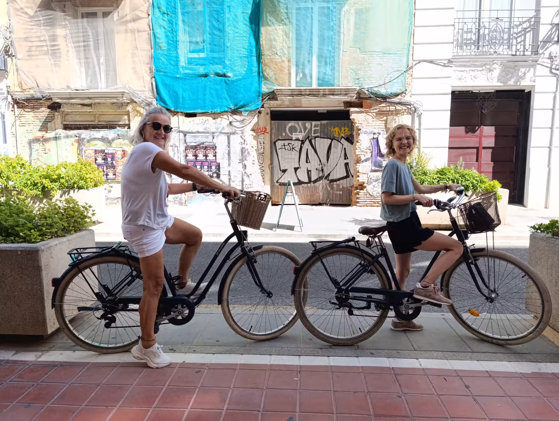 Two women on Dutch omafiets city bikes with baskets, pausing on a sunny street during bike rental