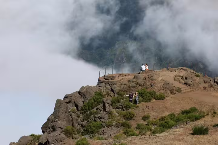 Travelers admire stunning views from a rocky peak in East Madeira, surrounded by lush greenery and misty clouds on a shore excursion.