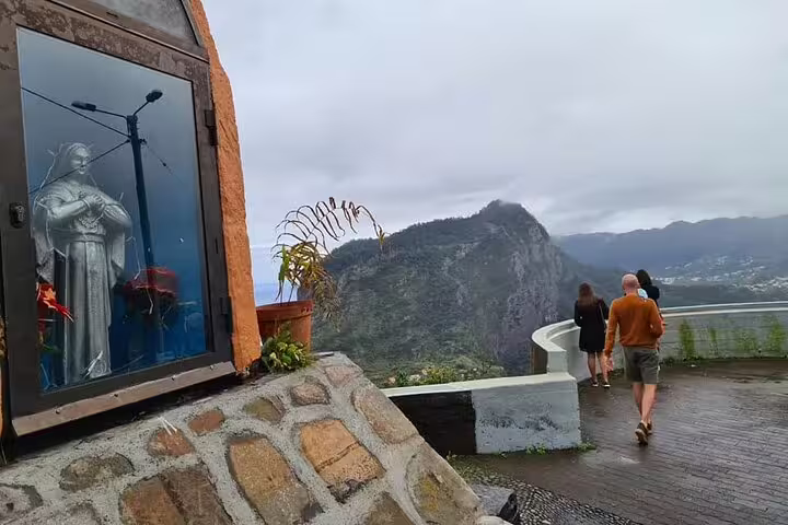 Tourists enjoy panoramic views from a scenic Madeira lookout with a nearby religious statue and lush mountains.