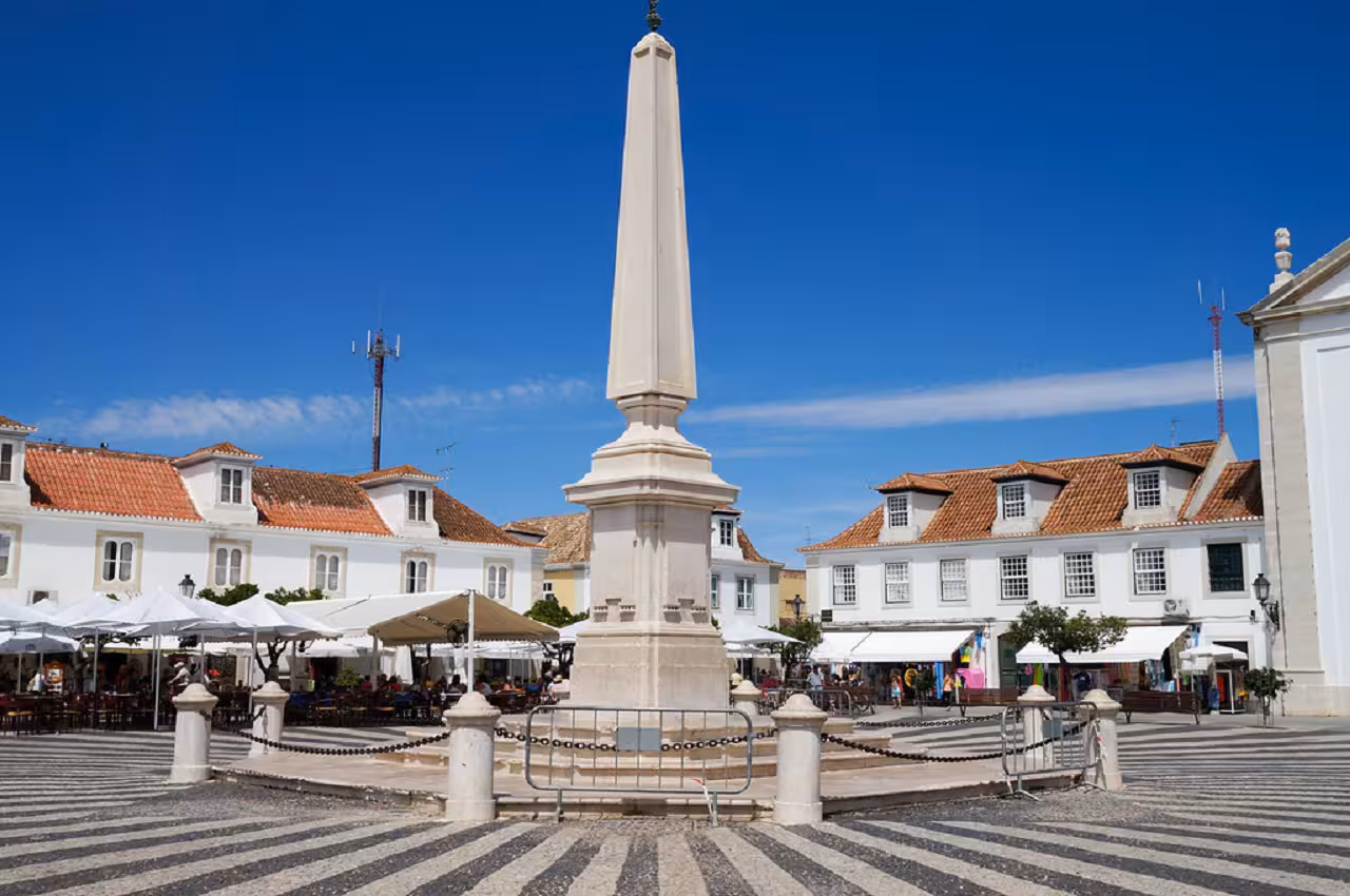 Central obelisk monument on patterned cobblestone square surrounded by cafés in a traditional Eastern Algarve town