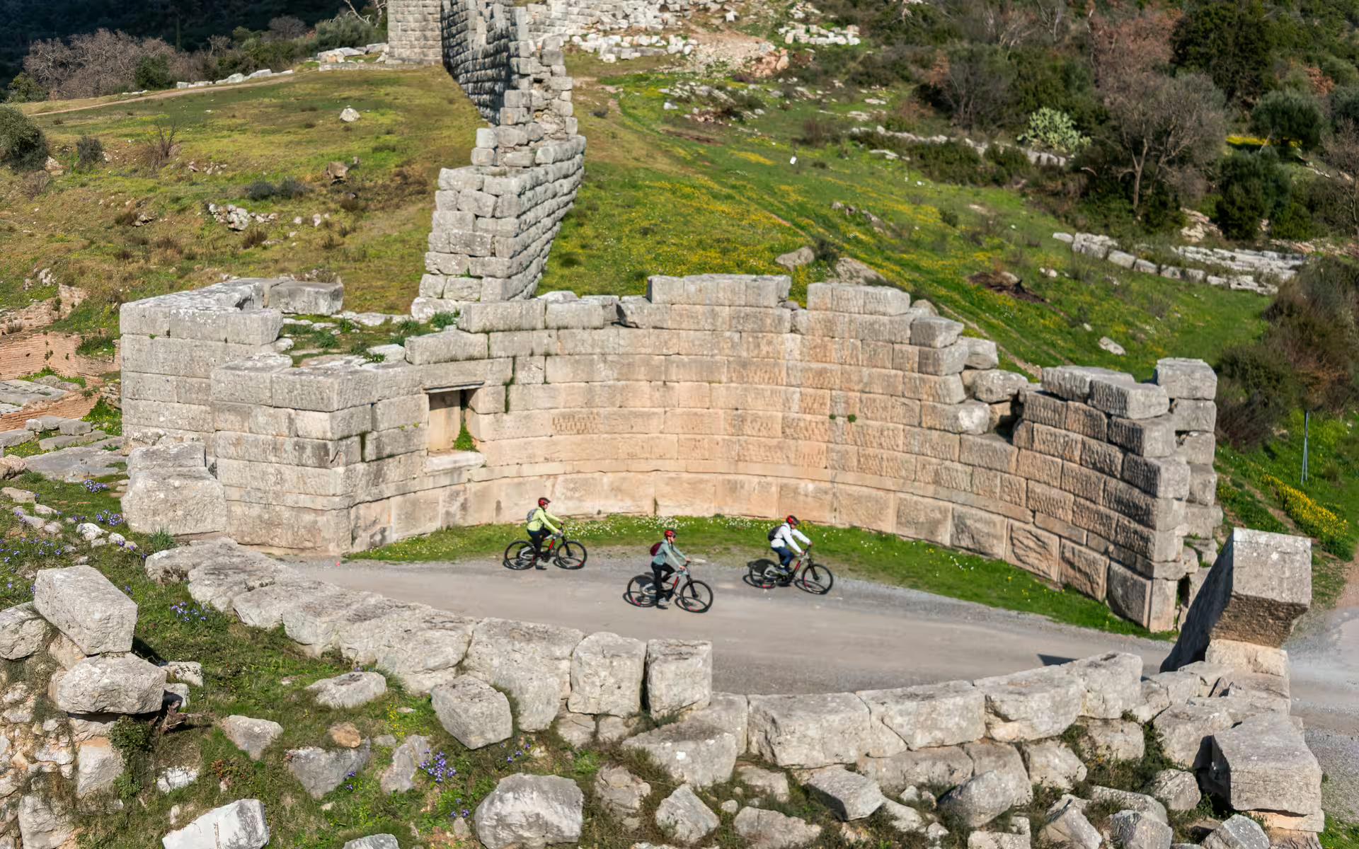 Group riding e-bikes past the ancient stone gate and curved walls of Ancient Messene archaeological site, Greece