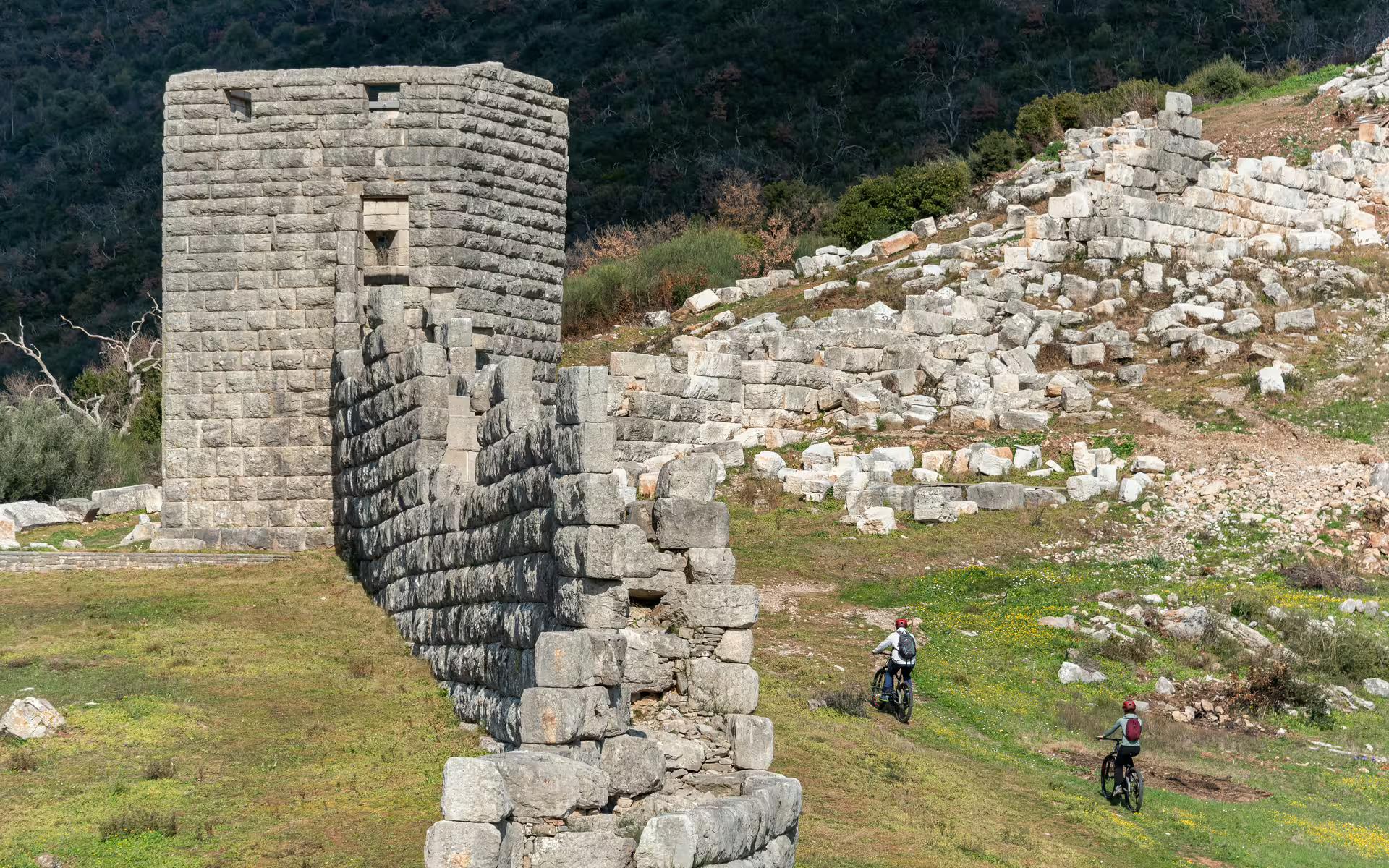 E-bike cyclists follow the Ancient Messene Way beside massive city walls and tower ruins in Messinia, Greece