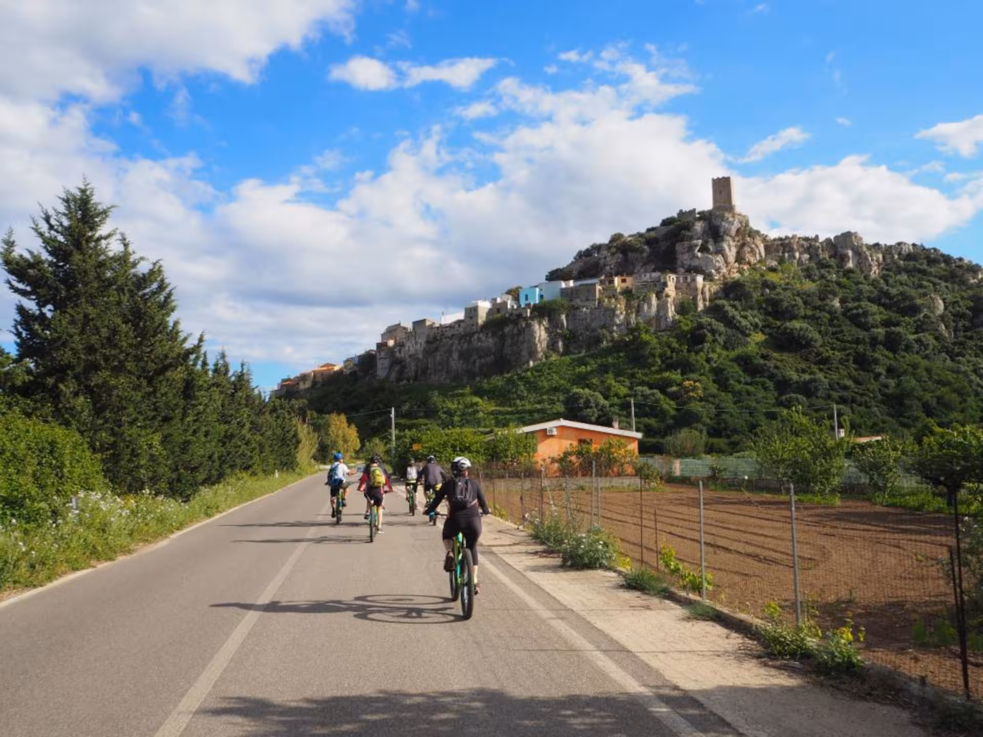Group of e-bikers riding along a picturesque road with a historic hilltop village in Sardinia.