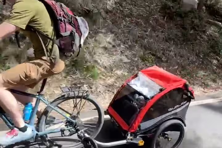 Cyclist with backpack pedals e-bike towing red trailer on scenic Sintra backroad.