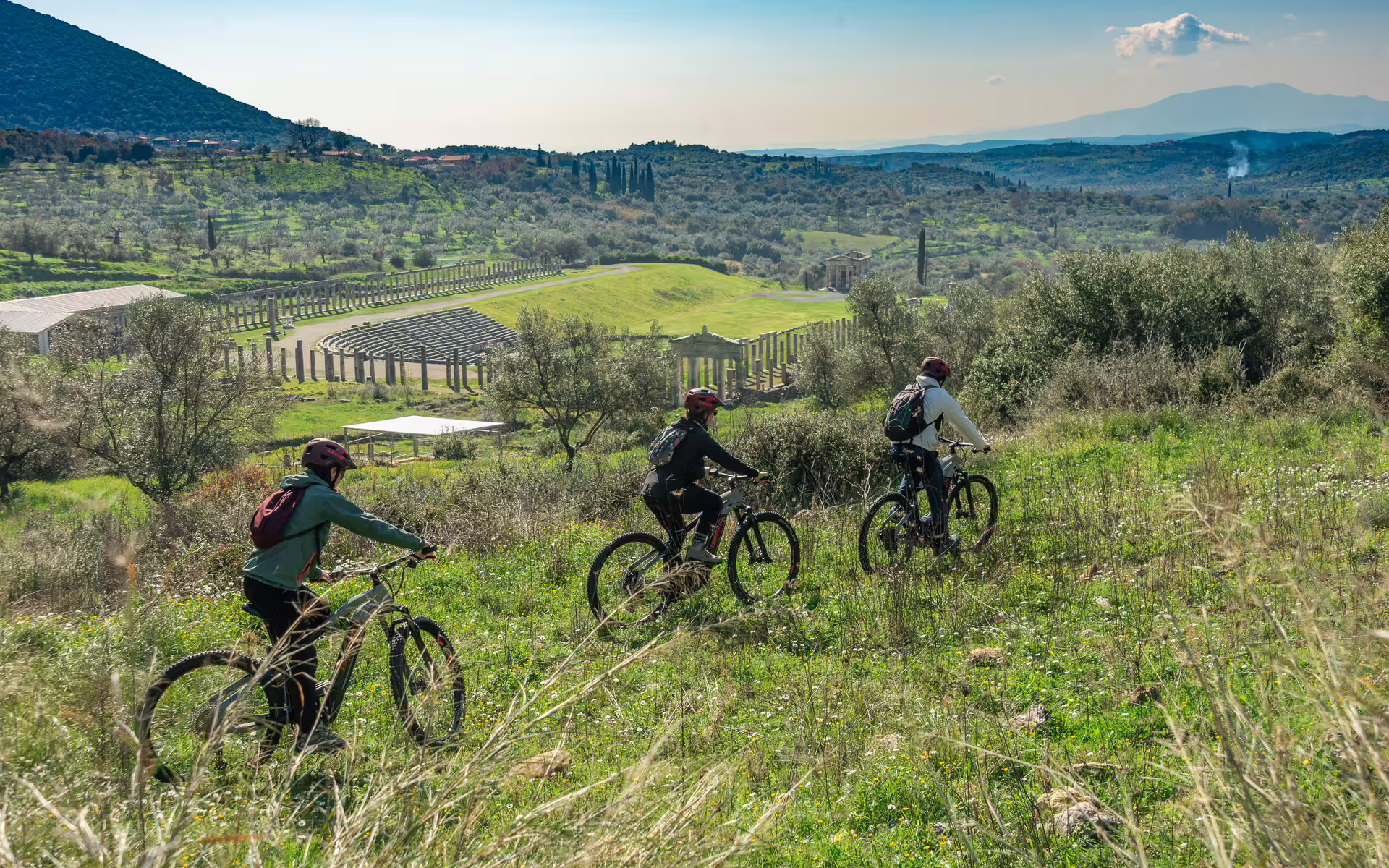 Guided e-bike tour riders above Ancient Messene ruins, cycling through olive groves with stadium views in Greece