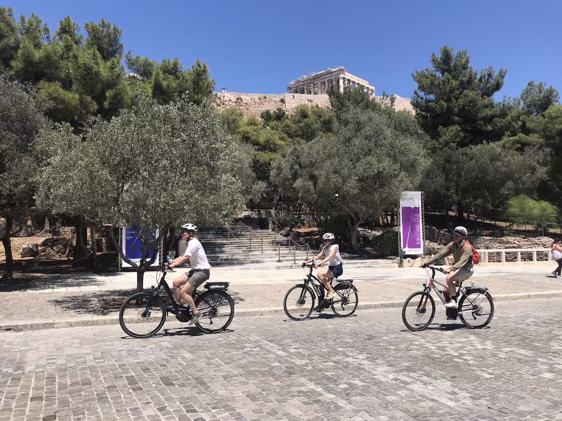 Cyclists on e-bikes ride past the Acropolis in Athens, enjoying a scenic tour under the clear blue sky.