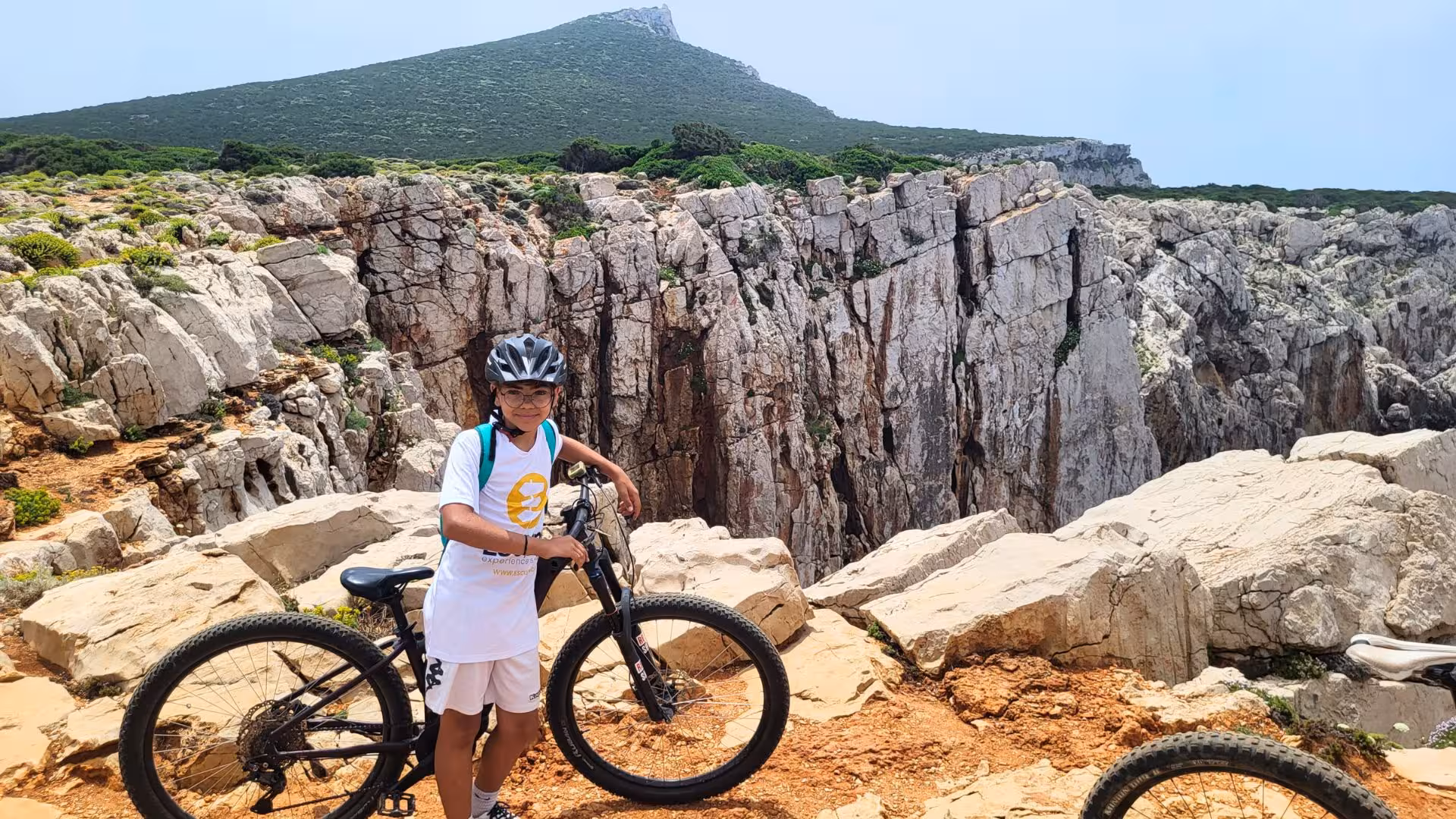 Young cyclist poses with e-bike on rocky terrain overlooking dramatic cliffs in Porto Conte Park, Alghero.