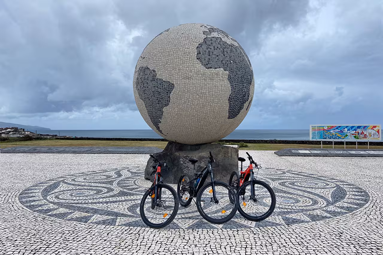 Three e-bikes parked before a giant globe sculpture on the oceanfront promenade during a guided Ribeira Grande cycling tour