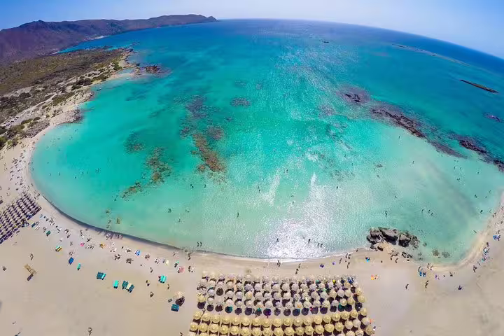 Aerial view of Elafonisi Beach with vibrant turquoise waters and pink sands, highlighting sunbathers and umbrellas on a private tour.