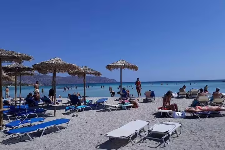 Visitors relax on sun loungers under straw umbrellas at Elafonisi Beach with its famous pink sands and clear blue waters.