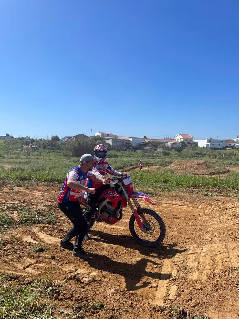 Instructor guides enduro rider during a practical training session on a dirt track in Carregado, Lisboa, under clear blue skies.