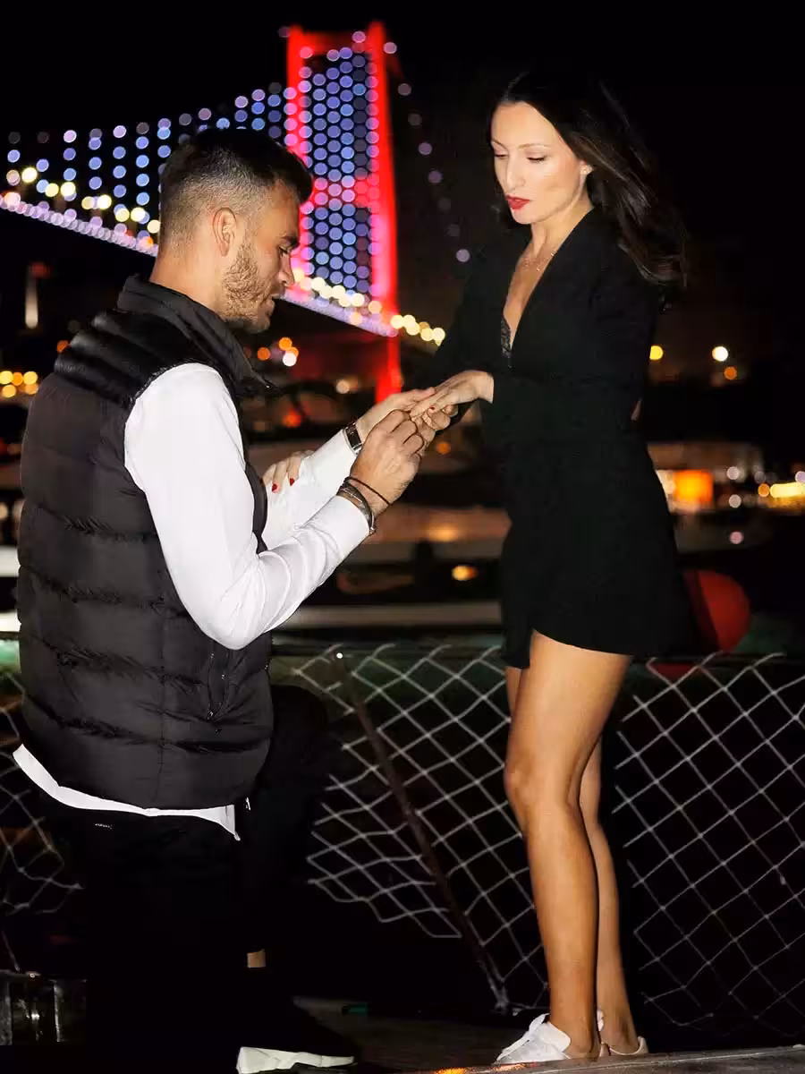 Man placing engagement ring on partner’s hand on a private yacht with Bosphorus Bridge lights, proposal setup