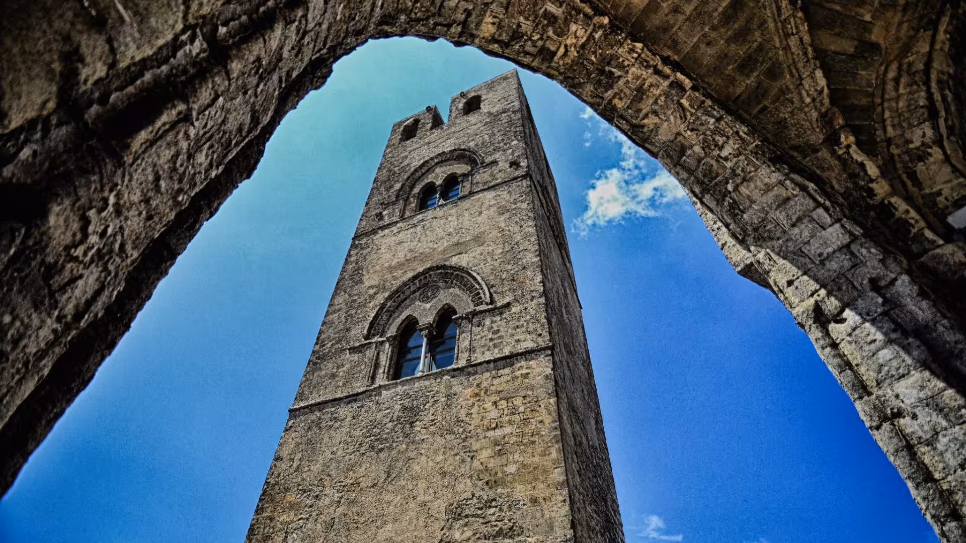 Majestic stone tower of Erice village framed by ancient archway against a vibrant blue sky.