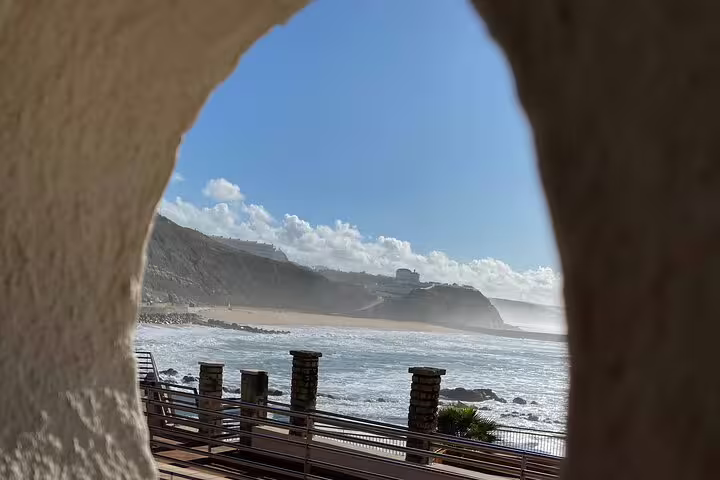 View through an arched stone opening framing Ericeira’s sandy bay and rugged coastal cliffs under a bright blue sky—highlighting the picturesque Ericeira coastline on the Tour to Ericeira and Mafra from Sintra.