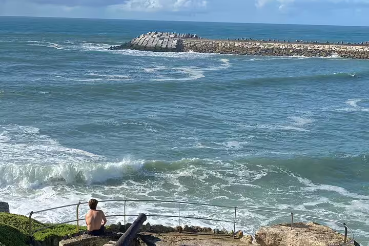 Traveler seated on the rocky coastal cliffs of Ericeira, gazing at the Atlantic Ocean’s rolling surf and a stone breakwater—showcasing the dramatic Ericeira coastline on the Tour to Ericeira and Mafra from Sintra.