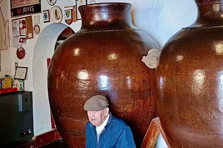 Elderly man in traditional attire sits beside large clay wine jars in a rustic Alentejo winery on the Evora wine tour.