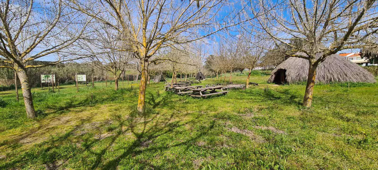 Reconstructed prehistoric huts and picnic area at Great Dolmen of Zambujeiro site in Evora, under a bright blue sky.