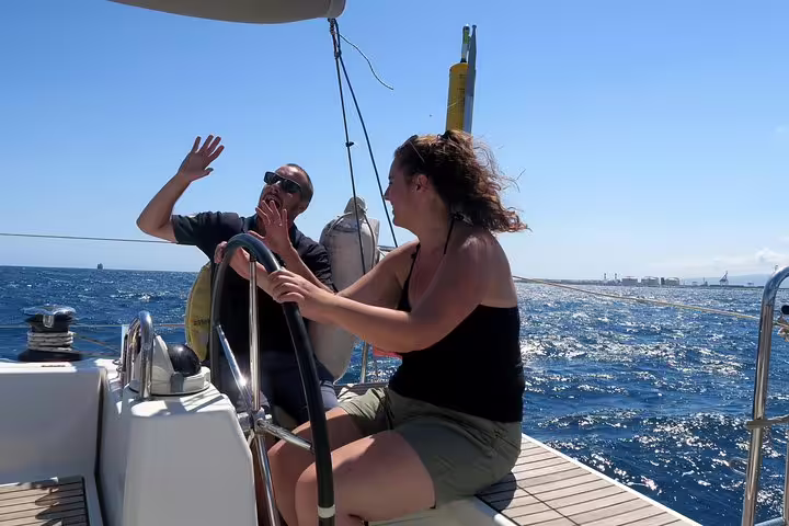 Two people enjoy steering a sailboat on a sunny day, capturing the essence of an exclusive sunset cruise from Barcelona.