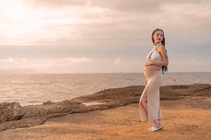 Expectant mother poses by the sea during a sunset photoshoot in Ammoudara, highlighting serene coastal beauty.