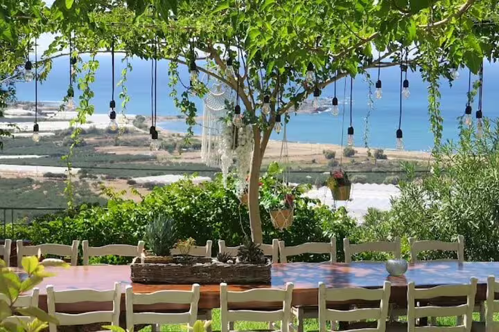 Charming outdoor dining setup under a leafy pergola overlooking the serene Falasarna Beach coastline.