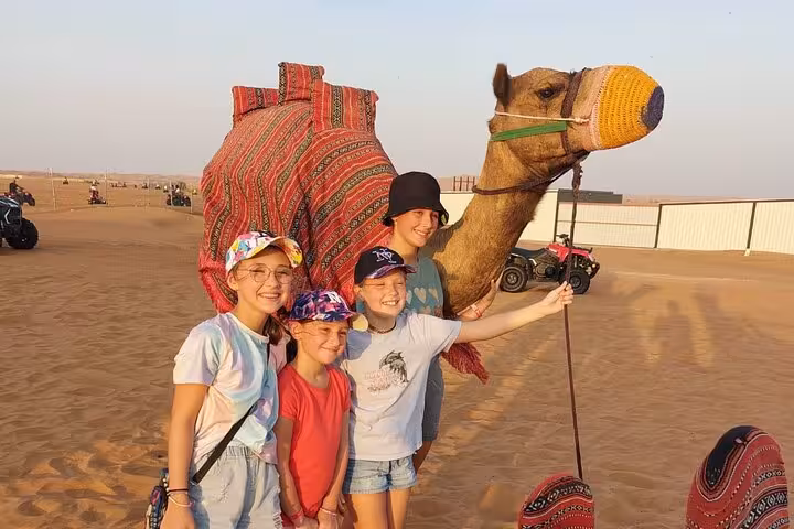 Family posing with camel in Dubai desert during Evening Desert Safari, with sand dunes and quad bikes