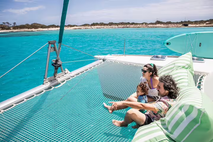 Family relaxing on a catamaran net with turquoise waters and scenic Formentera coastline, perfect for Ibiza small group trip.