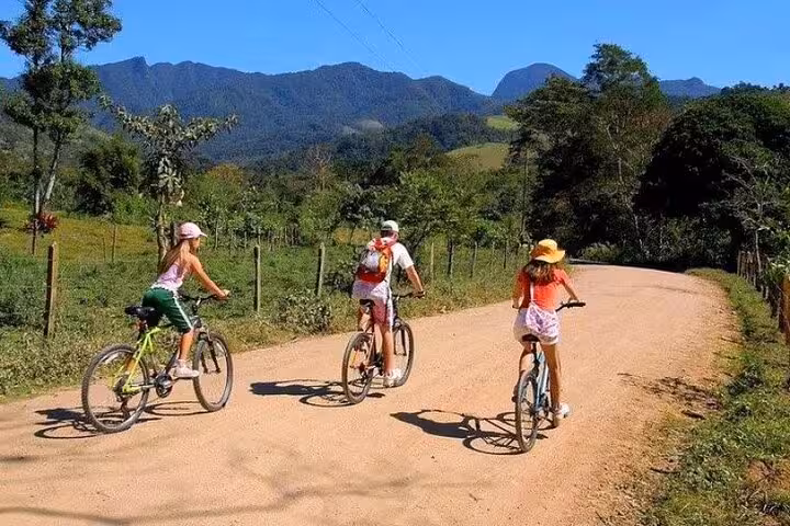 Family cycling on a scenic dirt road surrounded by mountains, showcasing Bike Rental by Paraty Tours.