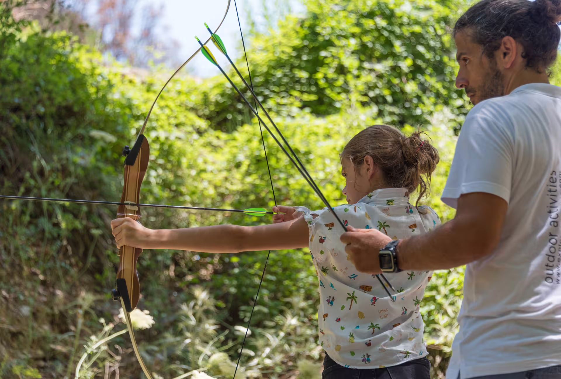 Family-friendly activity during Polylimnio Waterfalls hike, child learning archery in lush Greek forest