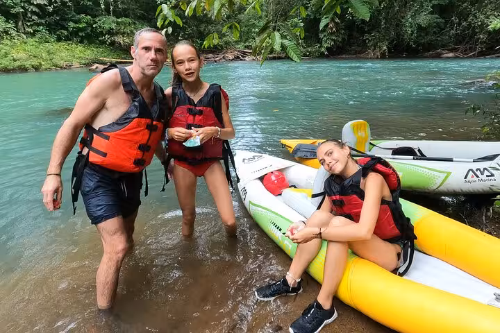 Family prepares for a kayak adventure, standing by the turquoise waters of Río Celeste in vibrant life jackets.