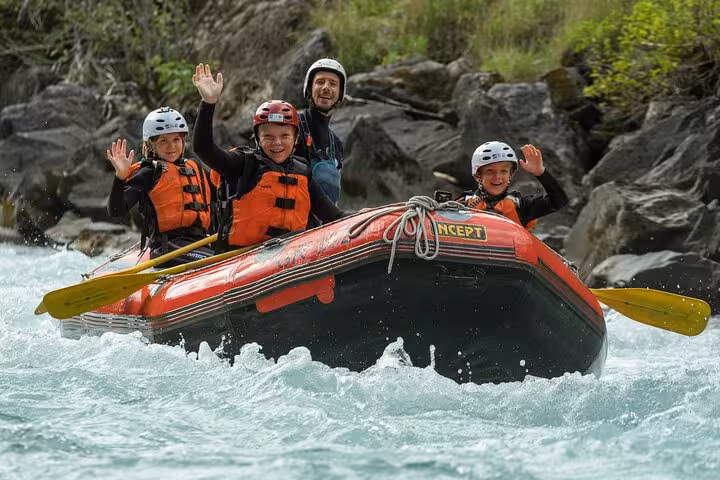 Smiling family waving during a fun rafting trip in Engadin, navigating calm waters with rocky landscapes in the background.