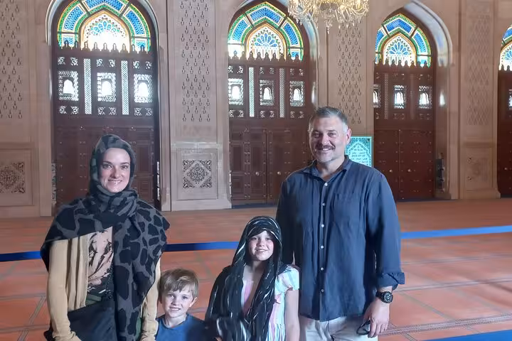 Family dressed modestly inside Sultan Qaboos Grand Mosque, admiring ornate interiors on Muscat half-day tour