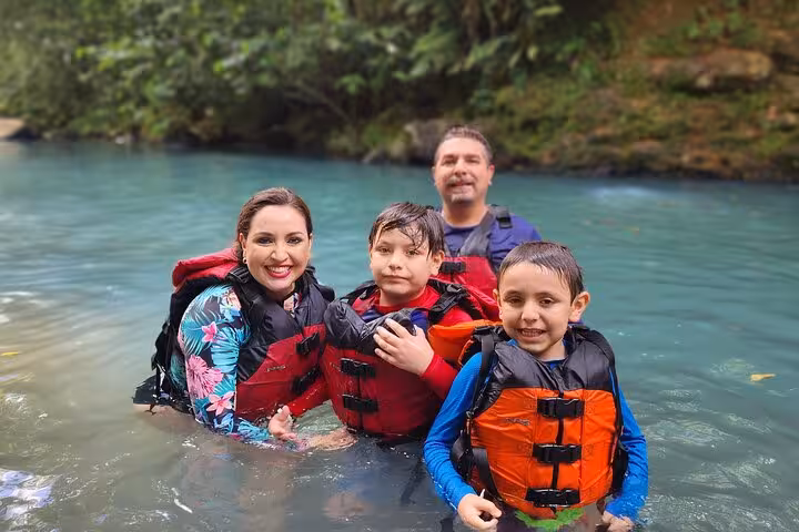 Happy family in life jackets standing in the clear waters of the Celeste River, surrounded by lush jungle scenery.