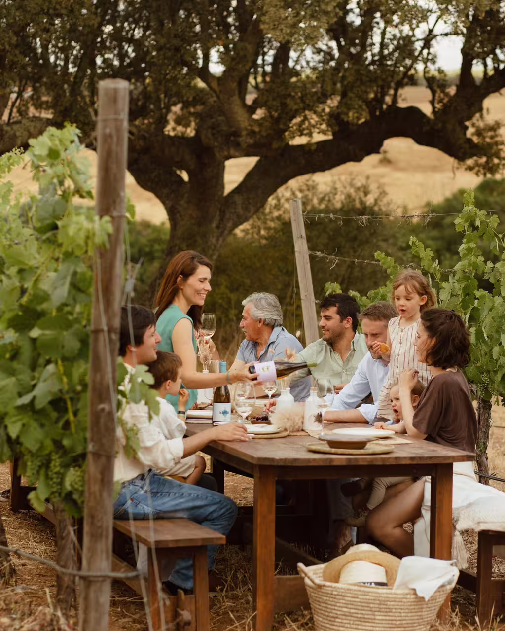 Family enjoying wine tasting tour outdoors in vineyard setting, with rustic table and lush grapevines under sunny skies.
