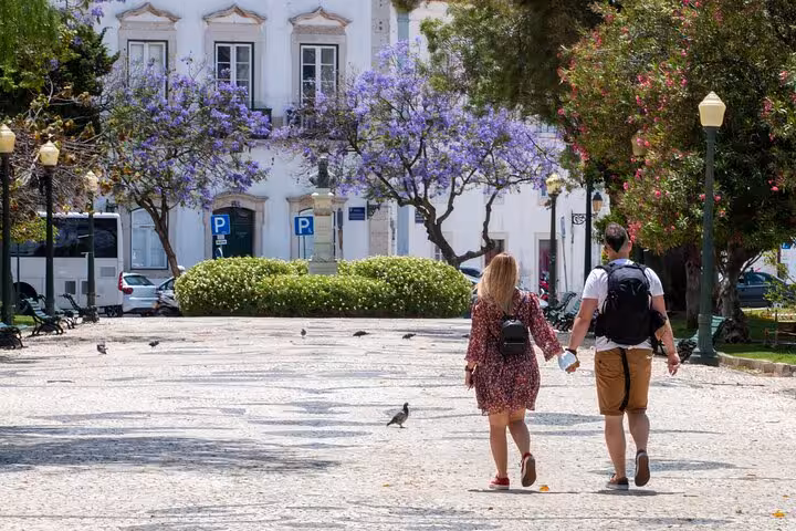 Couple strolling in Faro's charming square with blooming jacaranda trees on a sunny day, perfect for a city walk tour.