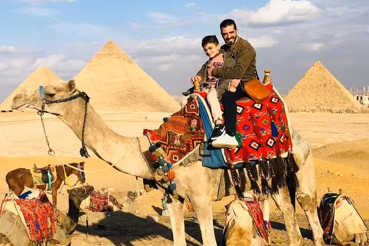 Father and son on decorated camel with the Giza Pyramids behind, private Cairo tour with Sphinx visit and lunch
