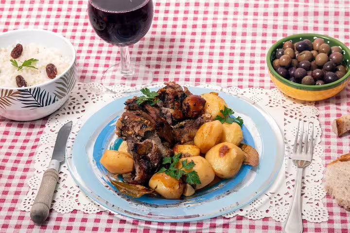 Traditional Portuguese meal with roast lamb, potatoes, olives, and red wine on a checkered tablecloth, highlighting Fatima and Coimbra tour flavors.