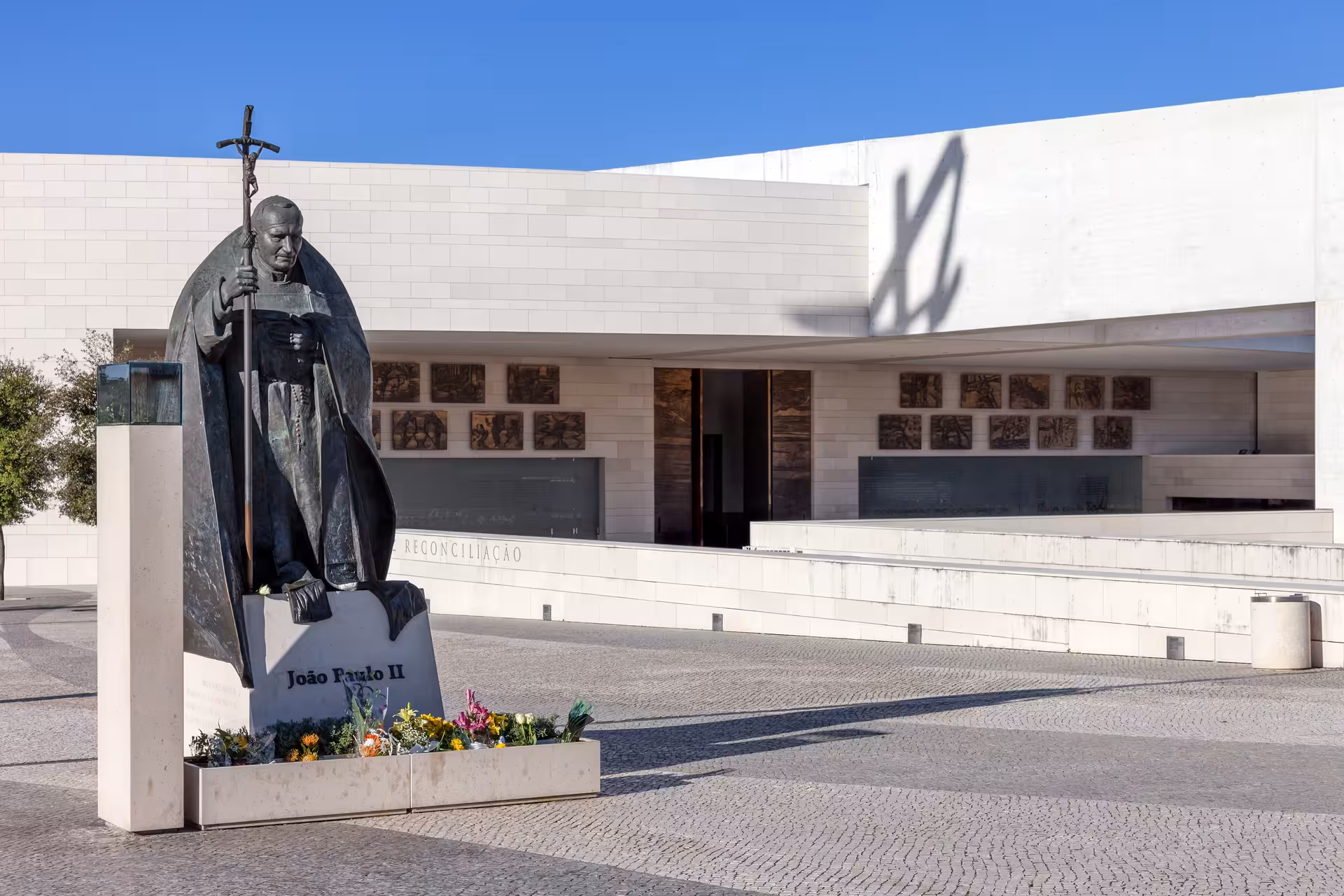 Statue of a religious figure outside the Fátima Reconciliation Chapel, surrounded by flowers under a clear blue sky.