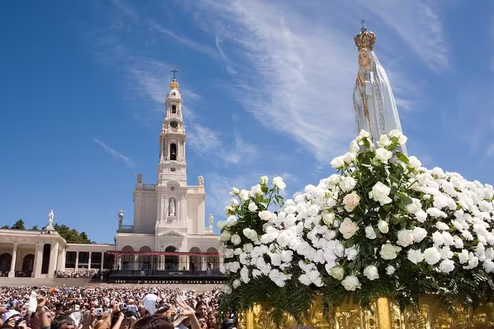 A vibrant crowd gathers at the Sanctuary of Fátima in Portugal, showcasing the iconic basilica and a statue adorned with white flowers.