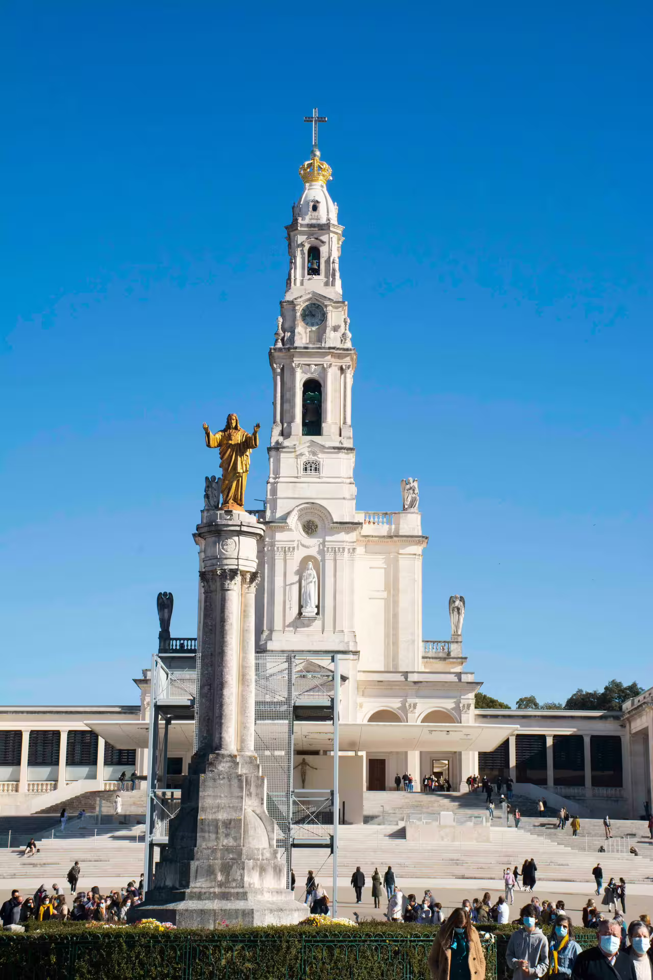The Sanctuary of Fátima under a clear blue sky, showcasing its iconic bell tower and religious statue, attracts numerous visitors.
