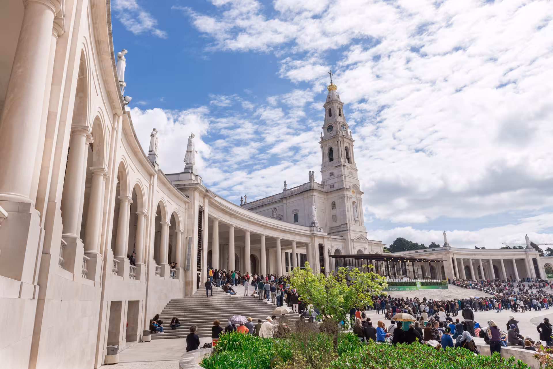 Crowds gather at the Sanctuary of Fátima, showcasing its impressive neoclassical architecture under a vibrant sky.
