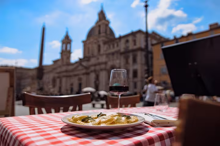 Plate of fettuccine pasta and wine on a checkered tablecloth with a scenic view of Rome city center architecture.