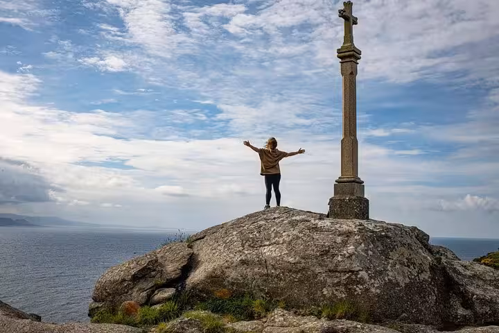 Traveler stands beside iconic cross on a rocky cliff overlooking the ocean at Finisterre, Costa da Morte tour highlight.