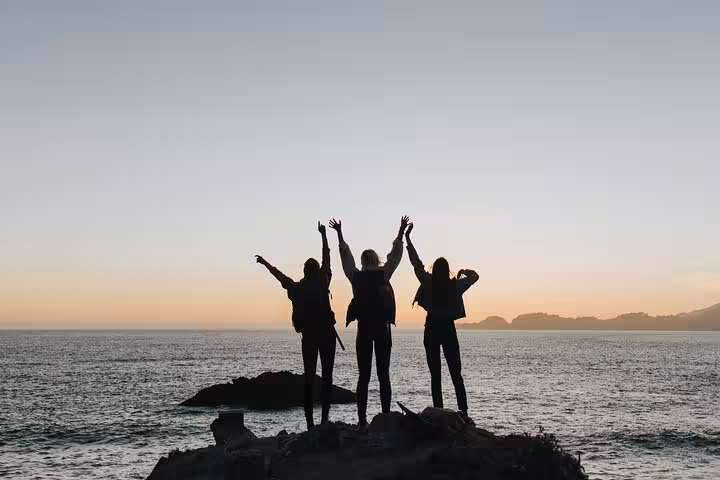 Three silhouetted people celebrate a scenic sunset over the ocean at Finisterre on a guided boat cruise excursion.
