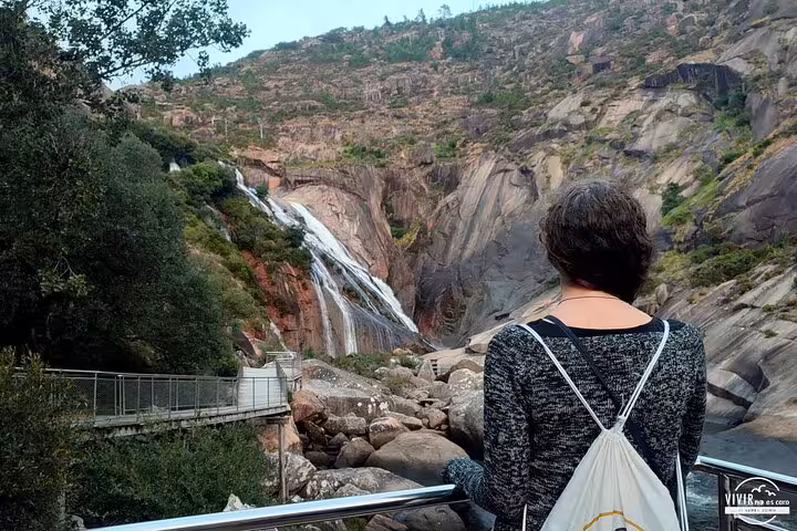 Woman gazing at the majestic waterfall in Finisterre, surrounded by lush greenery and rocky cliffs.