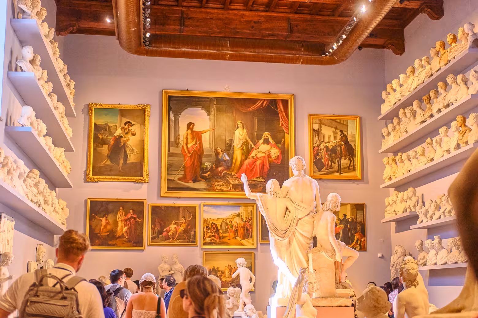 Visitors admire classical sculptures and paintings in a well-lit gallery room at the Accademia Gallery in Florence.