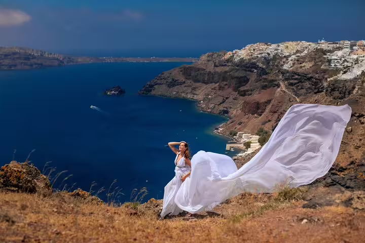 A woman in a sweeping white dress poses on a cliff overlooking the Aegean Sea in Santorini, perfect for a flying dress photo shoot.